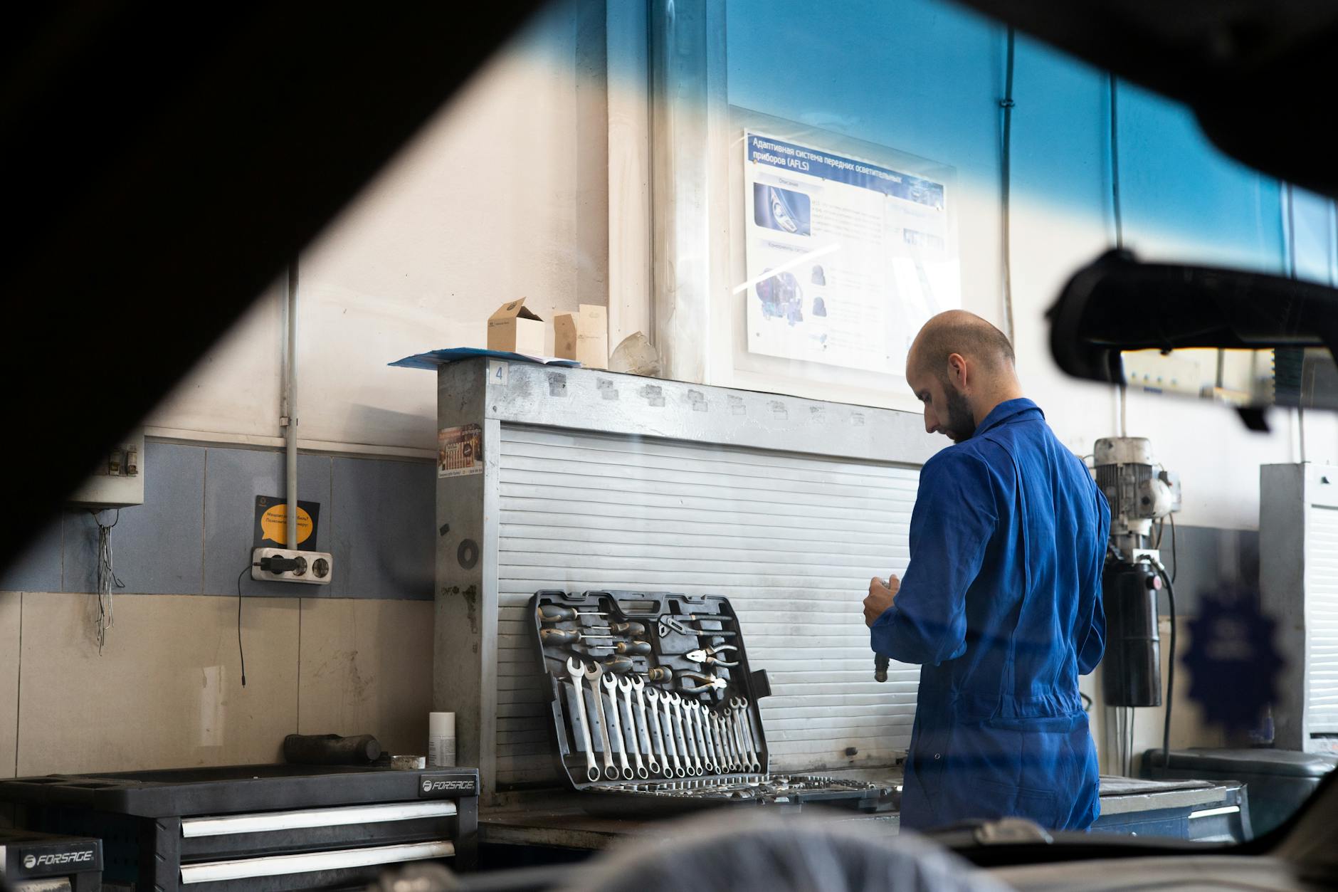 Bald mechanic in blue coveralls checking tools inside an automotive workshop.