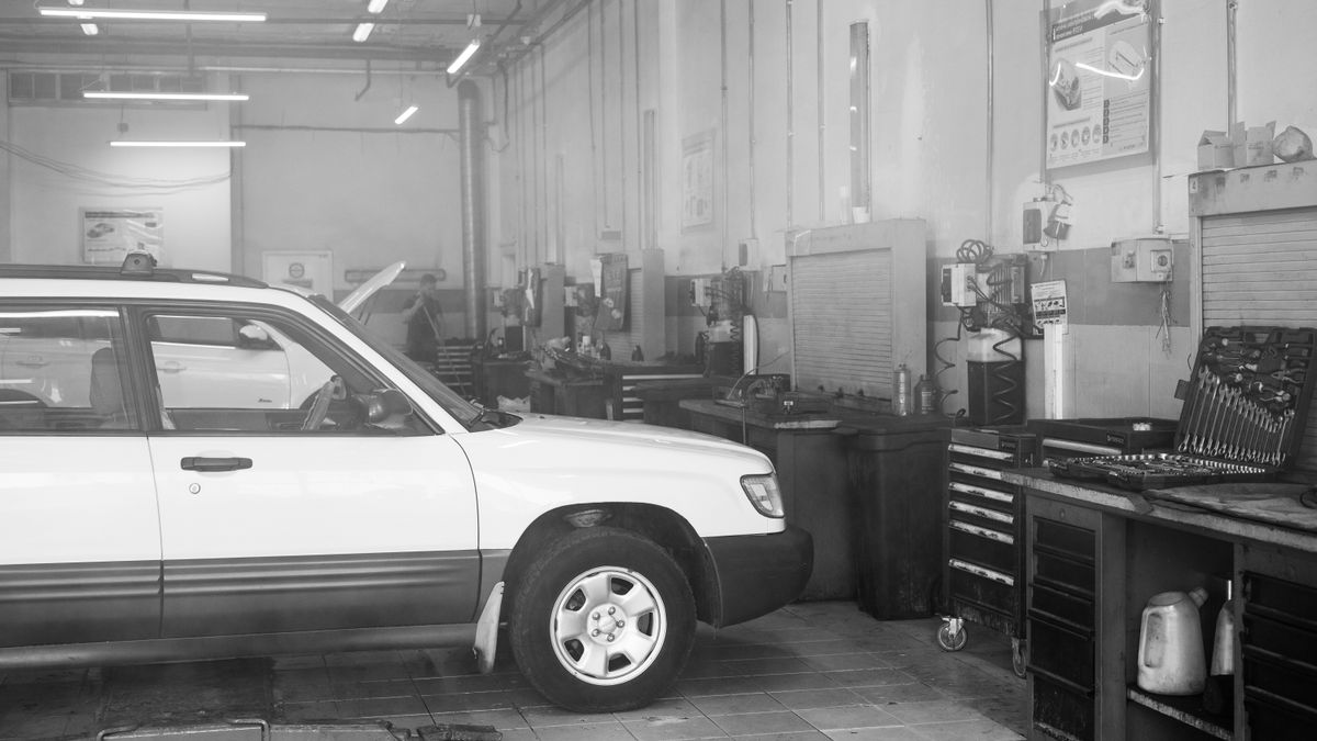 Black and white image of a car in a garage surrounded by tools and equipment.