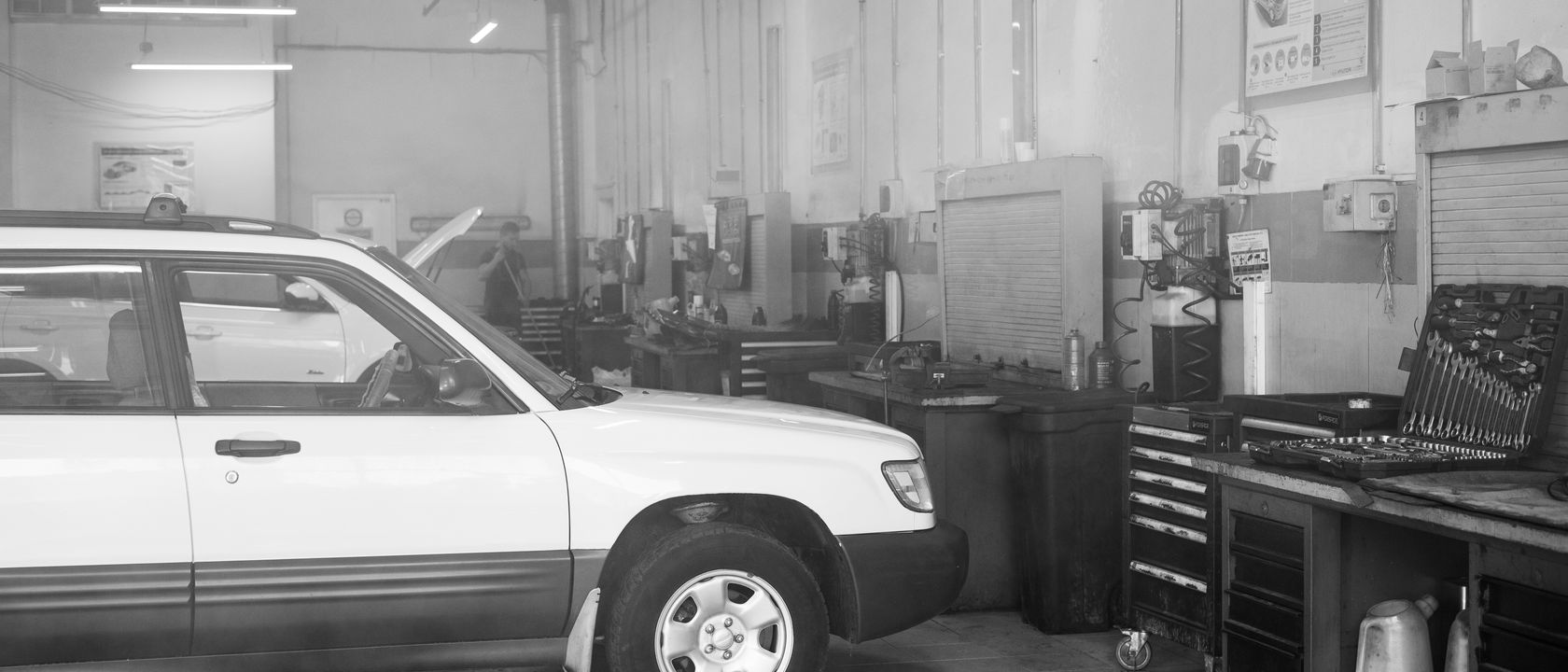 Black and white image of a car in a garage surrounded by tools and equipment.