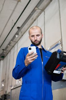 Mechanic in blue coveralls checking phone while holding oil container in a workshop.