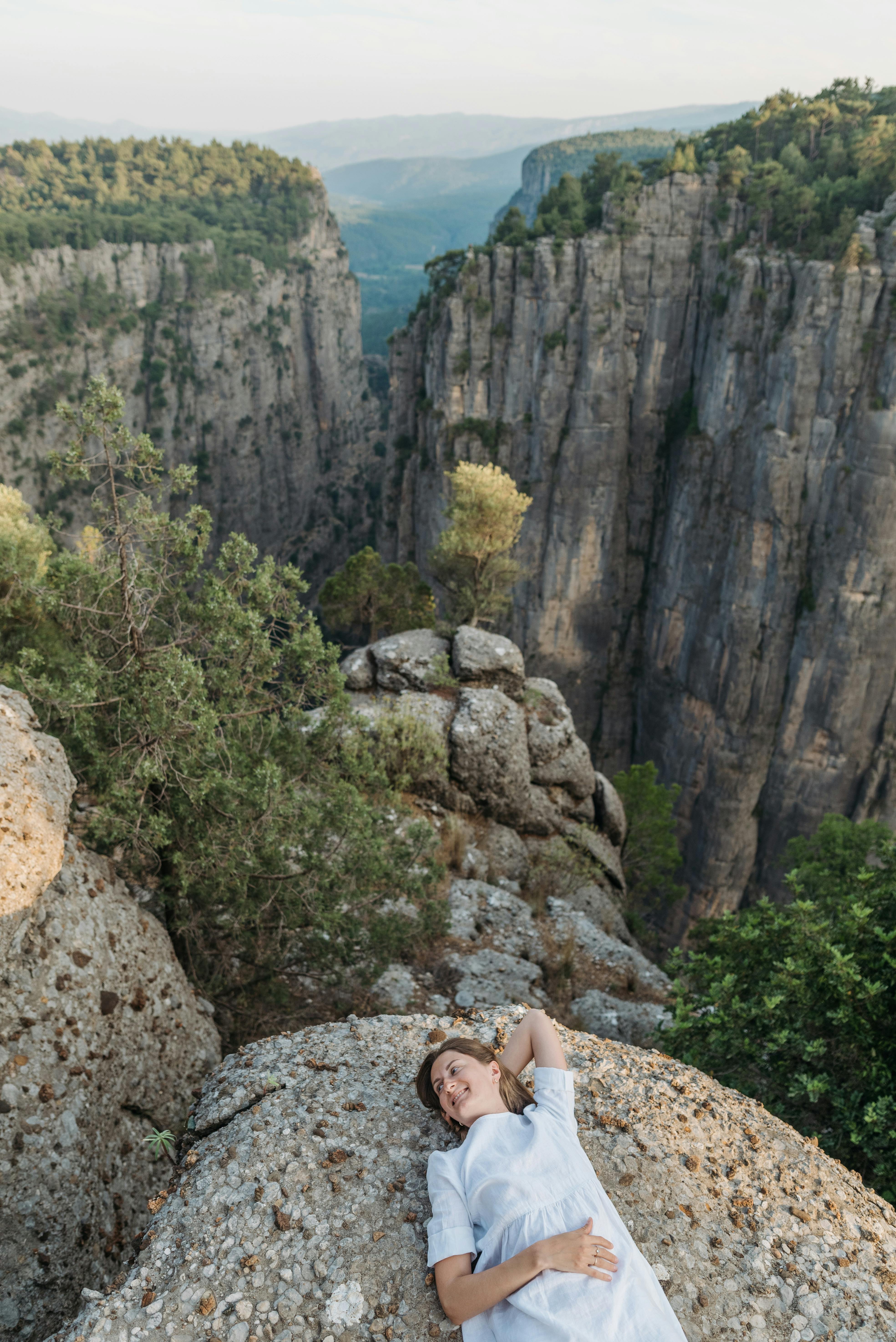 A woman relaxes on a clifftop overlooking dramatic rock formations and lush greenery.