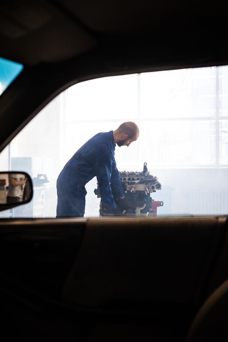 Mechanic working on an engine inside a garage, viewed through a car window.
