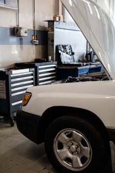 White car with hood open in a professional auto repair shop. Toolbox and equipment visible.