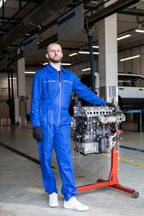 Man In Blue Zip Up Coverall Standing Beside A Car Motor Engine