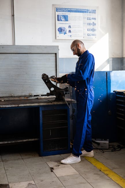 A focused mechanic in a blue jumpsuit performs work on a metal part in a well-organized garage workshop.