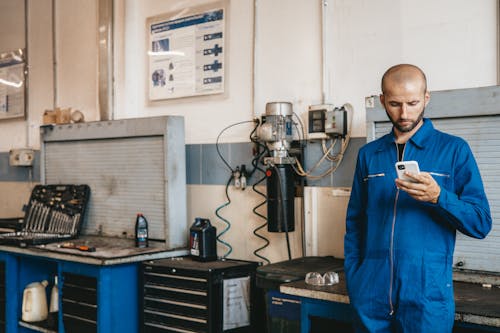 Free A Mechanic in Blue Coverall Using His Cellphone Inside  The Workshop Stock Photo