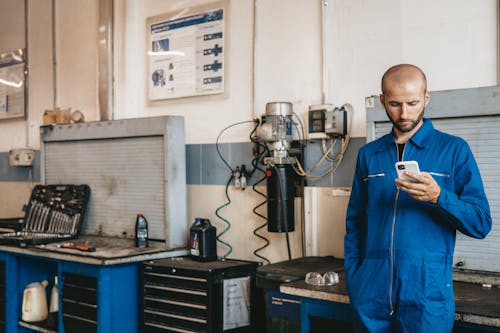 A Mechanic in Blue Coverall Using His Cellphone Inside  The Workshop