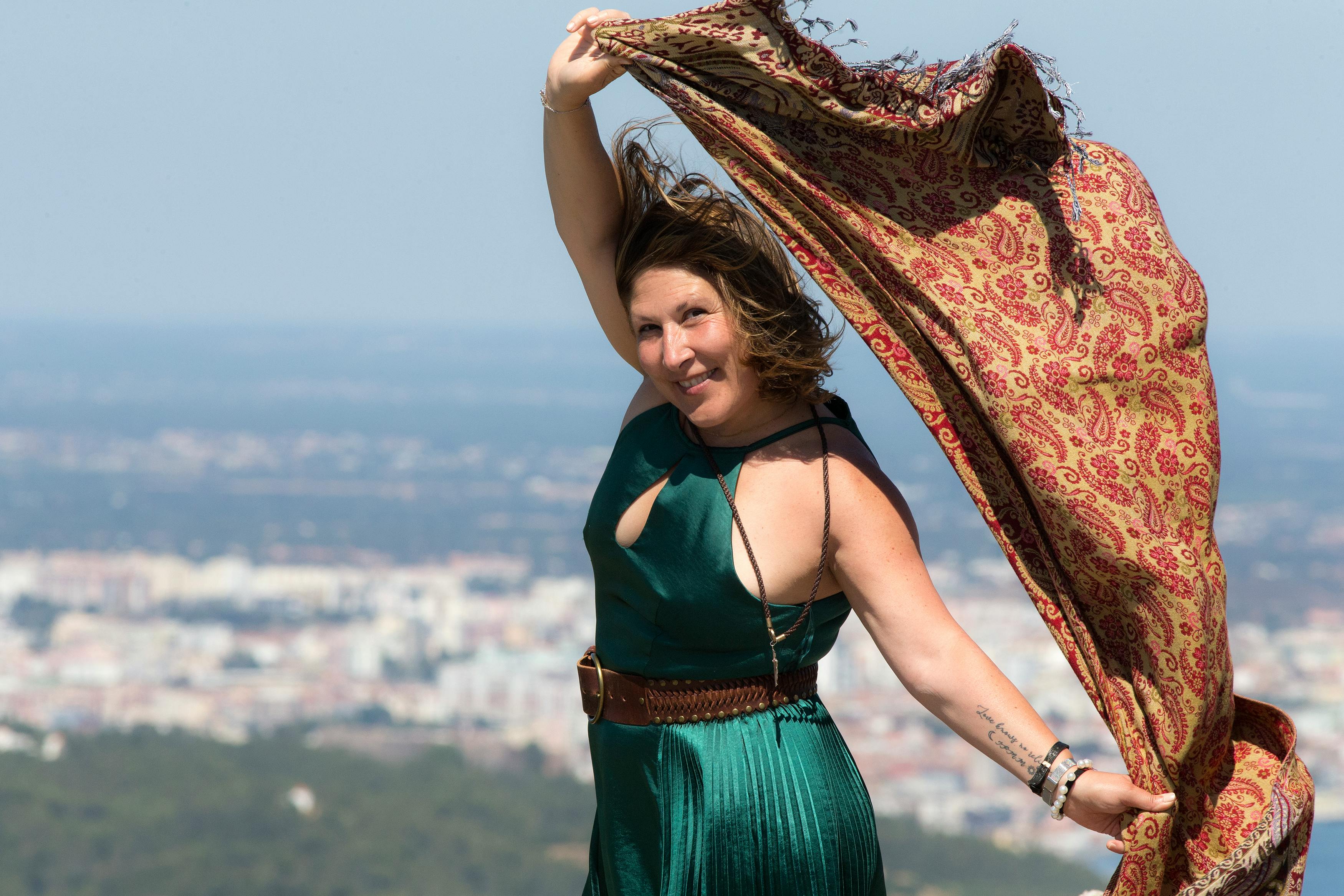An adult woman in a green dress joyfully posing with a scarf blowing in the wind.