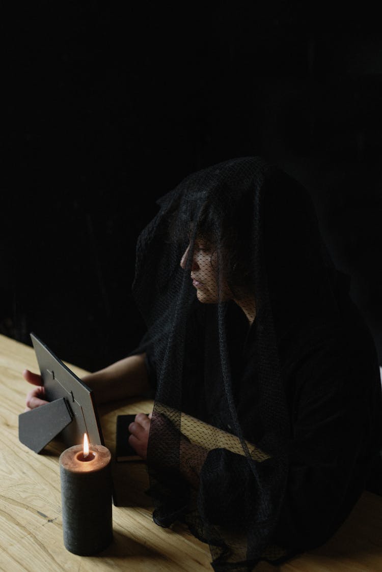 Black Image Of A Widow Sitting At A Desk With A Candle And Photograph