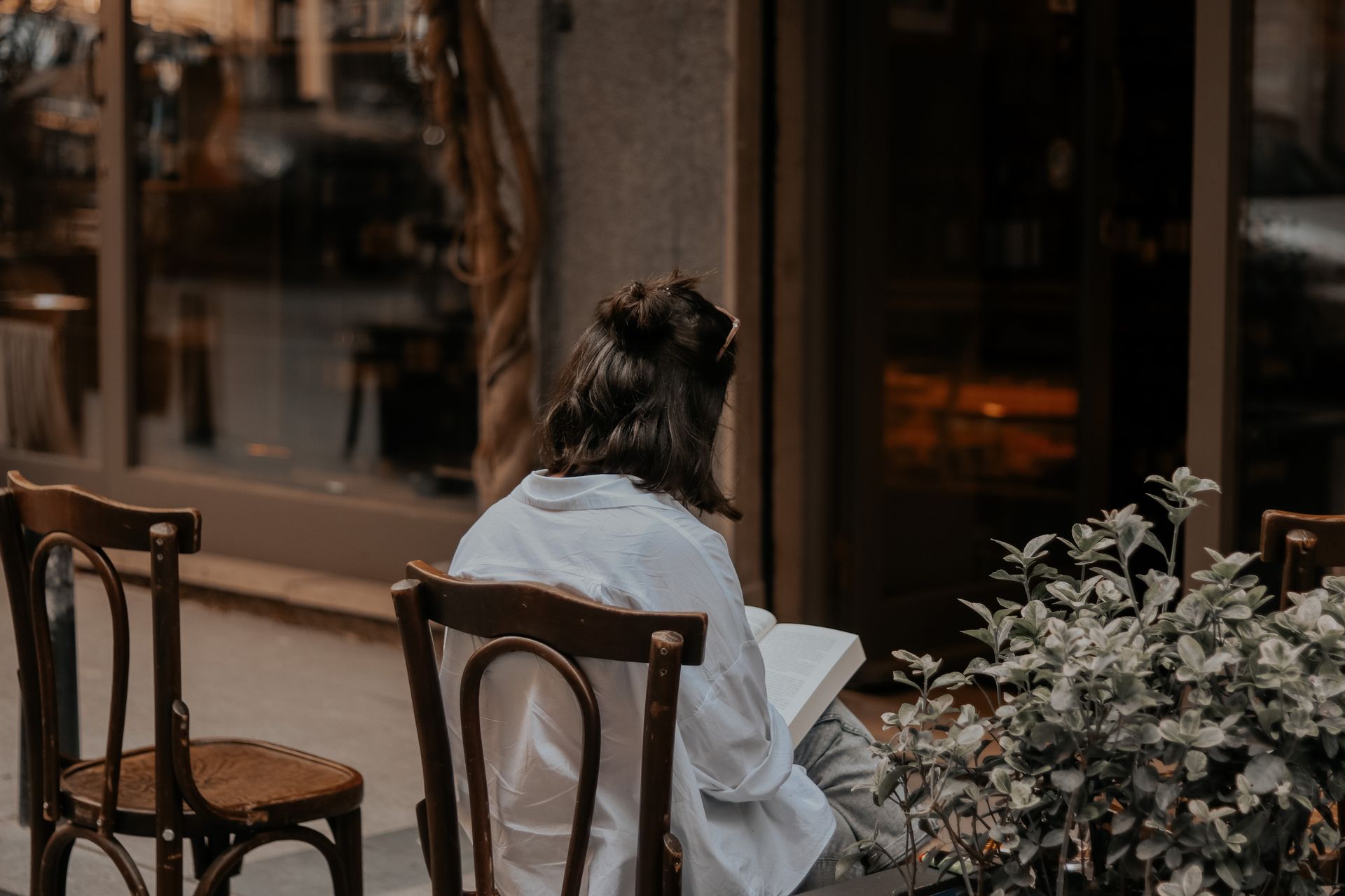 A woman enjoys reading a book outdoors at a quaint street cafe, surrounded by plants and wooden chairs.