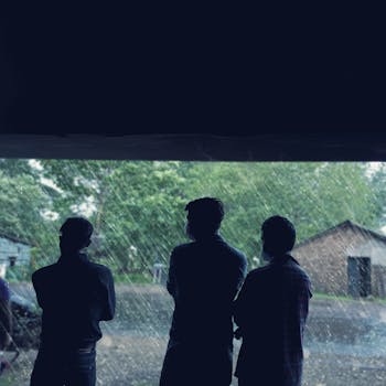 Three silhouette men stand sheltered indoors watching a rainy scene outdoors in India.