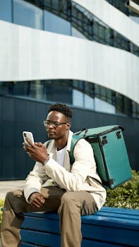 Modern food delivery person using smartphone while sitting on a city bench.
