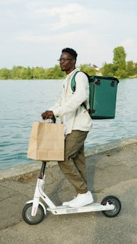 An urban delivery person rides an electric scooter carrying a backpack and paper bag by a river.