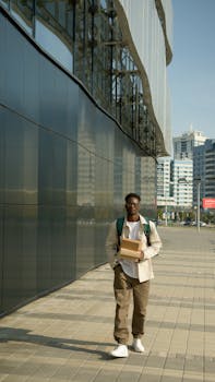 Delivery person walking on sidewalk with packages outside a modern building in urban setting.