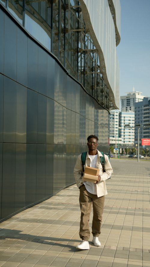 Free Delivery person walking on sidewalk with packages outside a modern building in urban setting. Stock Photo