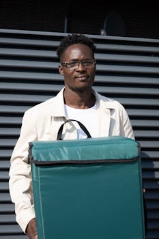 African American male delivery courier holding a green insulated bag in a sunny outdoor setting.