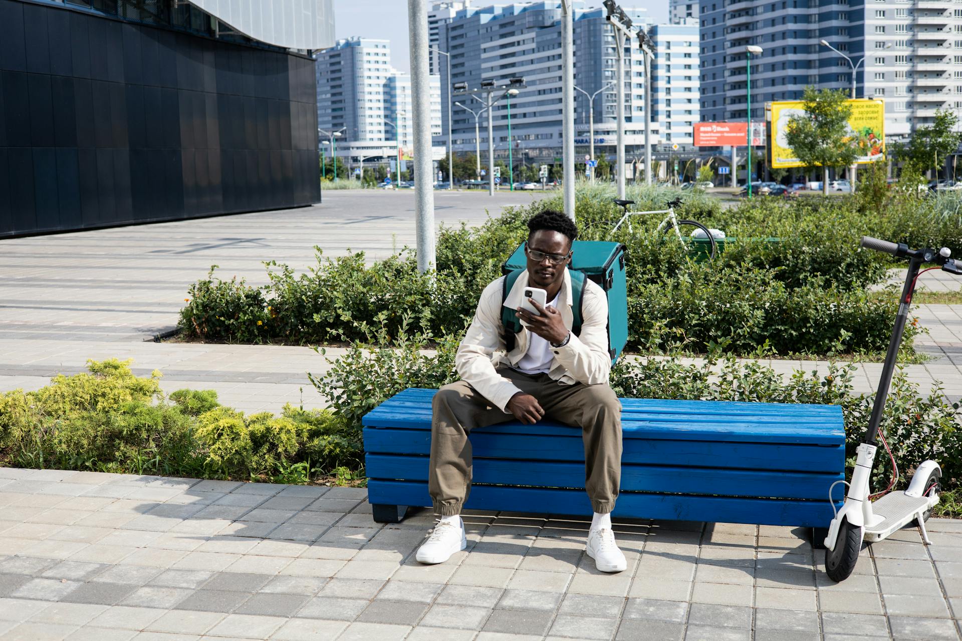Young delivery man on a break with electric scooter and thermal bag in modern cityscape.