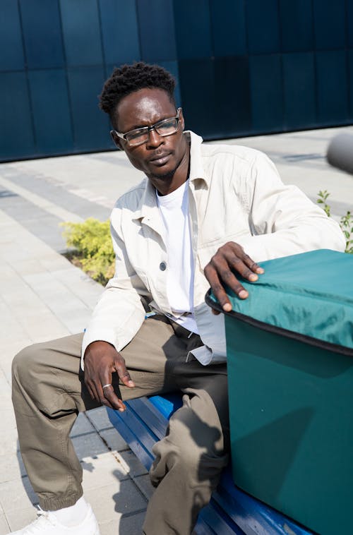 Man in White Dress Shirt Sitting on a Blue Bench Beside a Thermal Bag