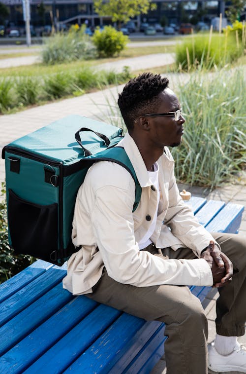 Free Man in White Dress Shirt and Brown Pants Sitting on Blue Wooden Bench Carrying a Thermal Bag Stock Photo