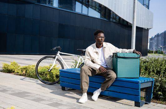 A delivery man rests on a bench outdoors with a bicycle and a thermal bag.
