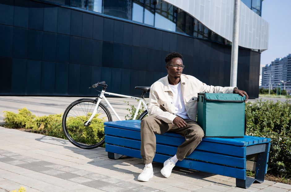 A delivery man rests on a bench outdoors with a bicycle and a thermal bag.