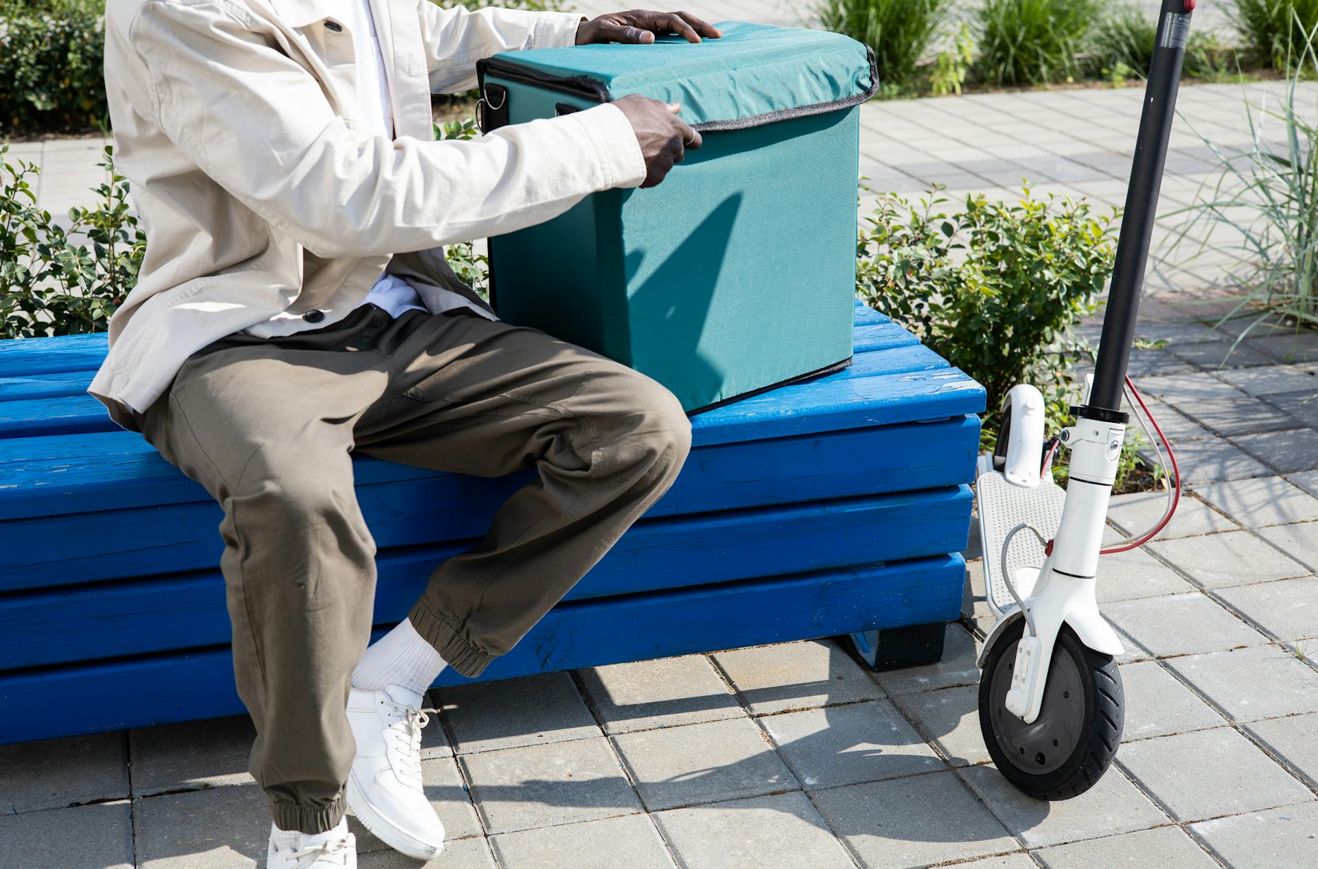 A delivery person with a thermal bag sitting on a blue bench next to an electric scooter.