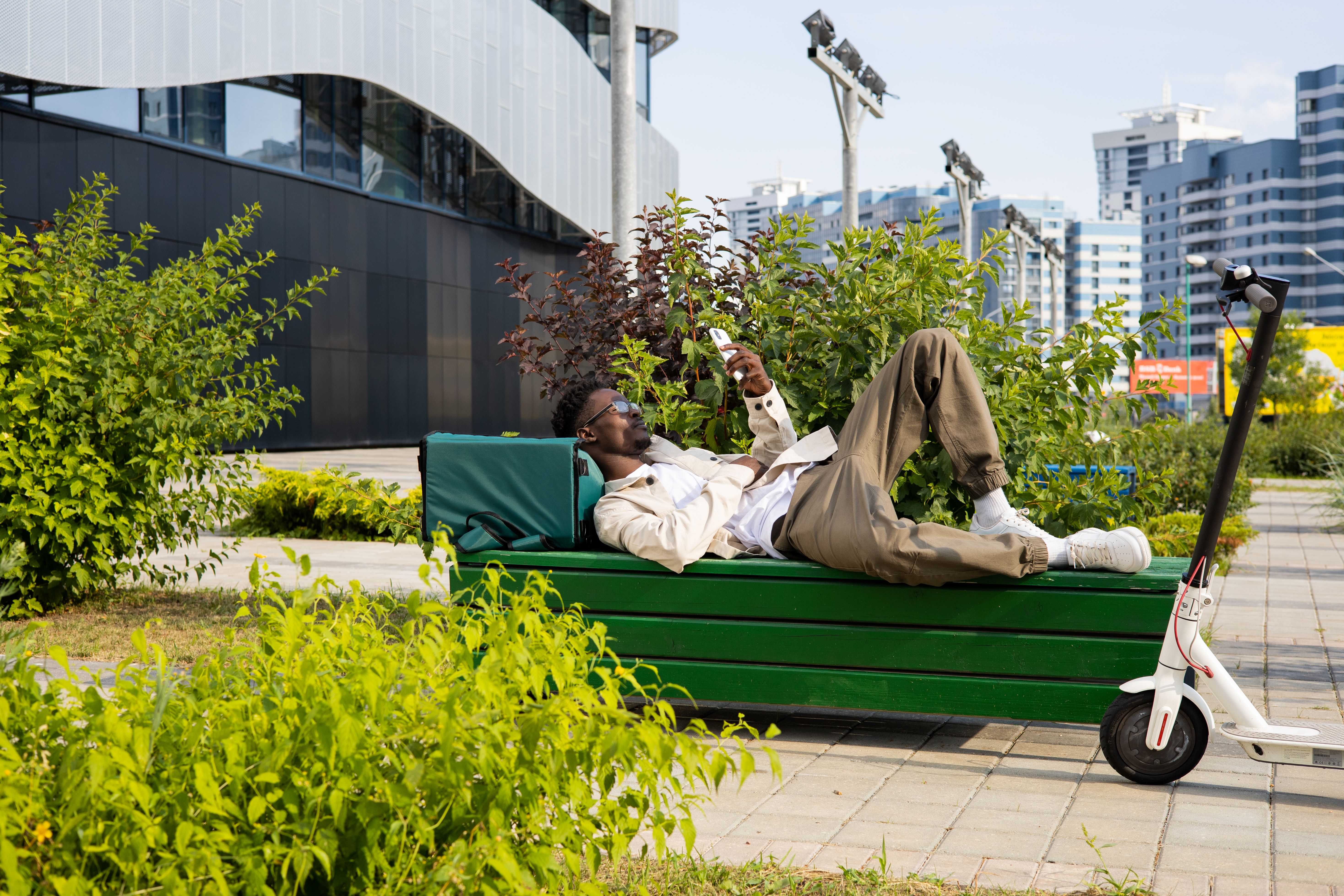 Man relaxing on park bench with smartphone next to electric scooter.