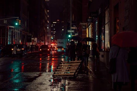 Rain-soaked city street scene at night with illuminated buildings and pedestrians under umbrellas.