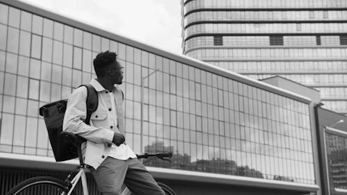 Grayscale Photo of a Man Sitting on a Bicycle while Looking at the Glass Building