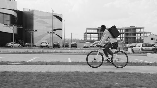 Free Black and white image of a deliveryman cycling on a city street with modern architecture. Stock Photo