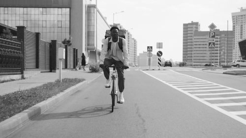 Free Black and white image of a courier cycling through city streets with buildings in the background. Stock Photo