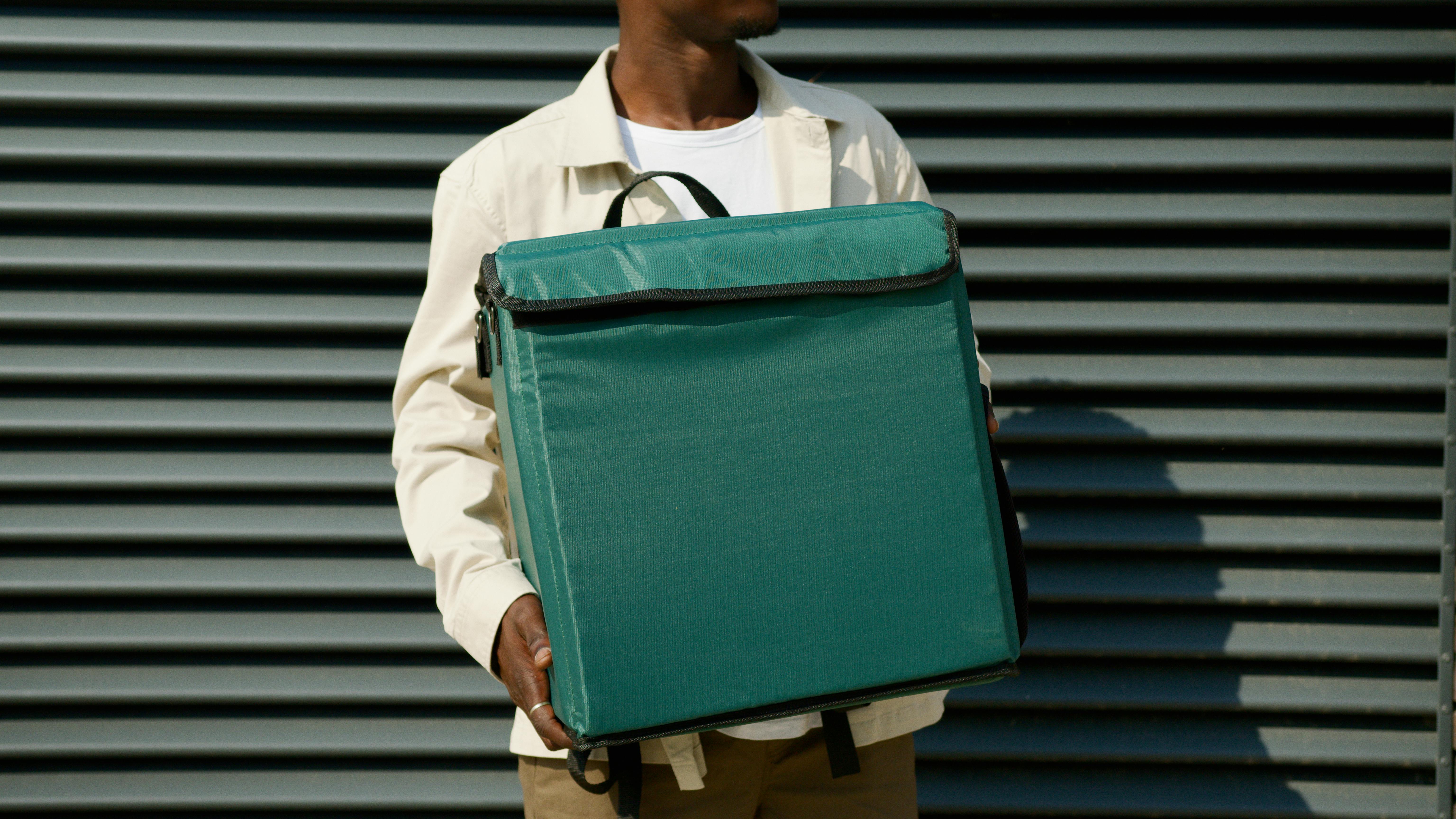 Delivery worker holding a large green insulated package outside a building with metal shutters.