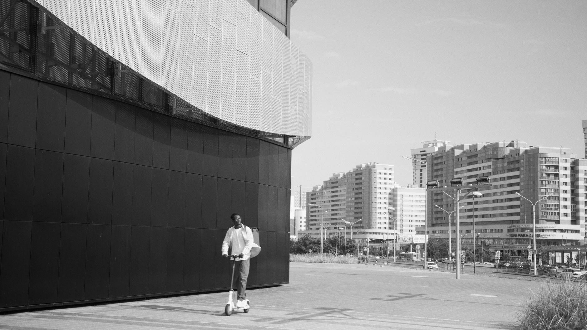 Black and white image of a man on an electric scooter amidst city buildings.
