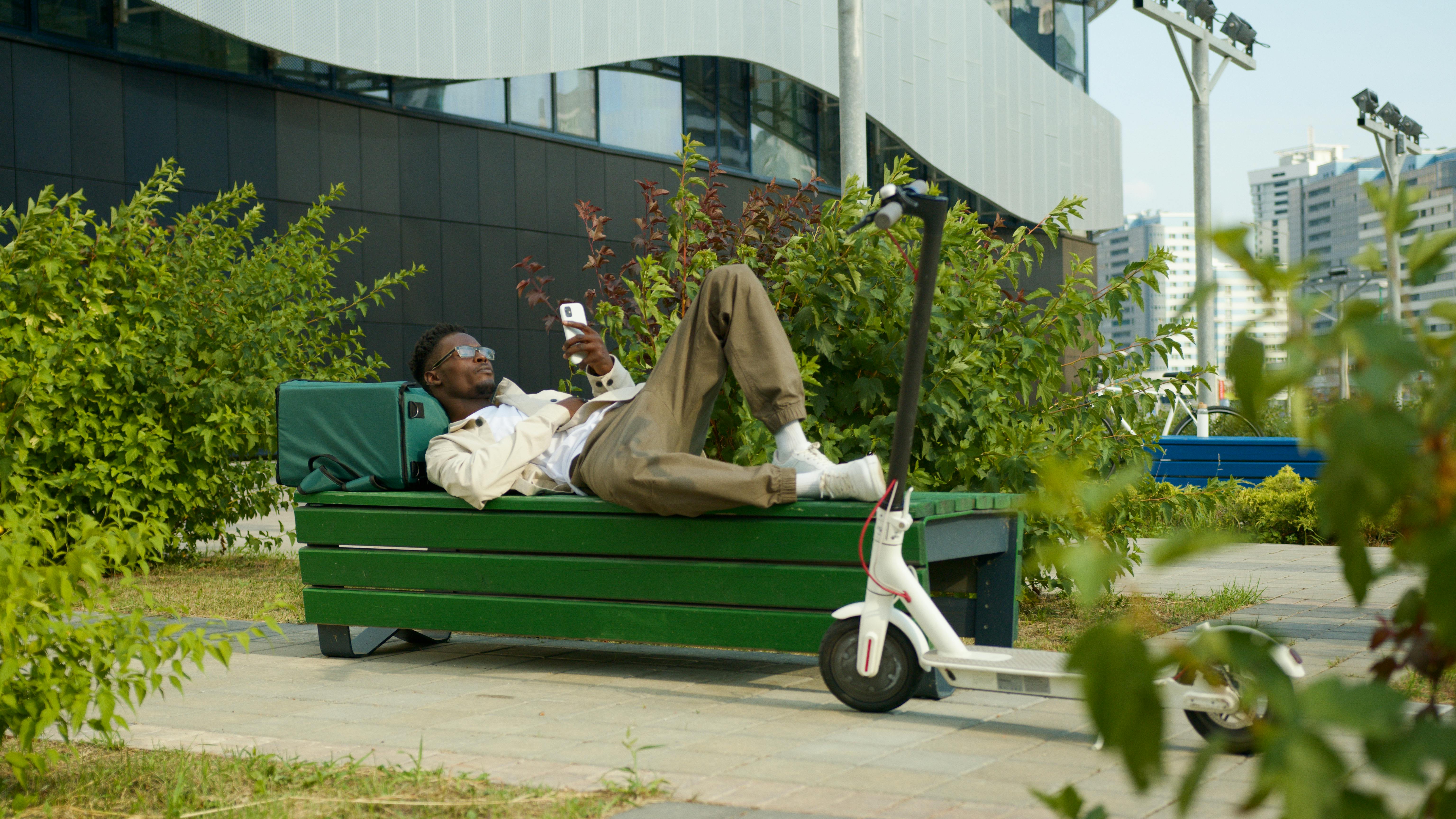 Man lounging on a bench using smartphone, electric scooter nearby.
