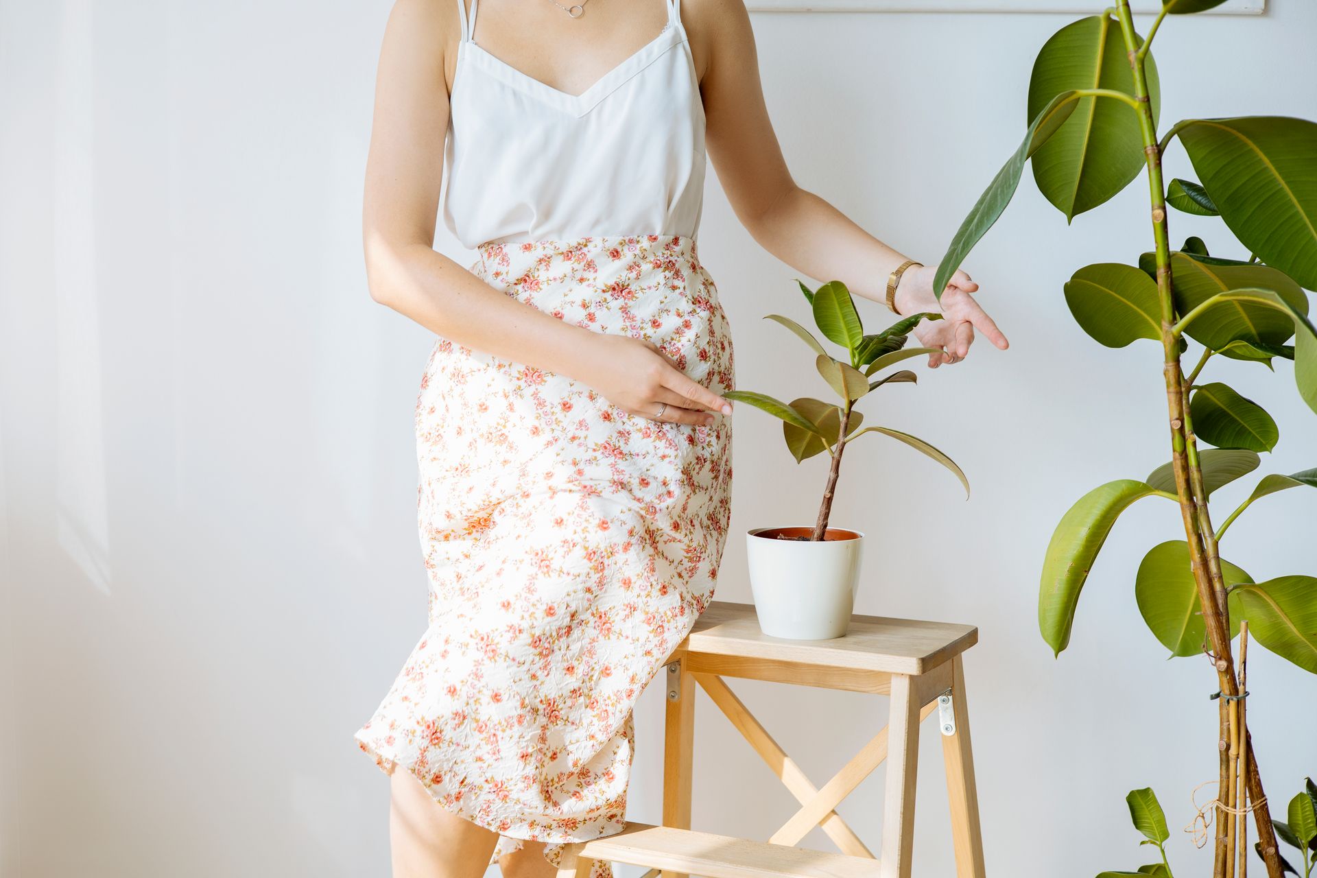 Woman gently cares for potted plants indoors, dressed in a floral skirt and white top.