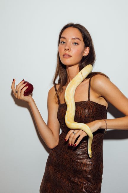 Fashionable woman in a stylish dress holds a red apple with a snake draped over her shoulders.