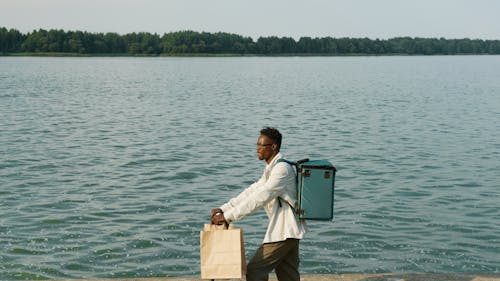 A Man Holding Brown Paper Bag with a Thermal Bag Standing Near Body of Water