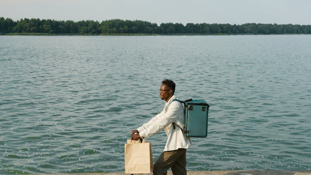 A man with a thermal backpack and paper bag walks along a lakeside, delivering food.