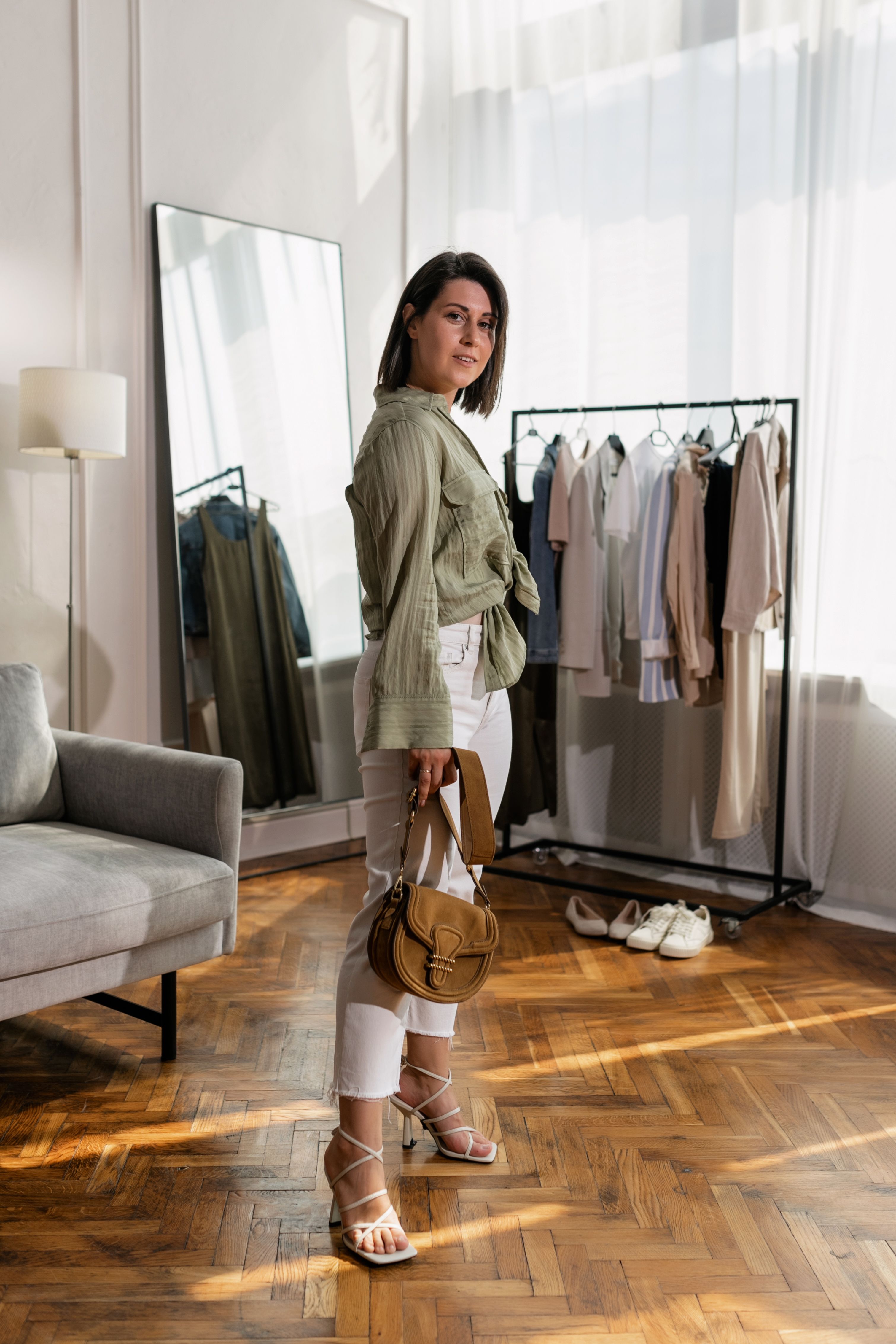 Woman styling a relaxed green shirt with white trousers and a brown shoulder bag in a bright wardrobe room
