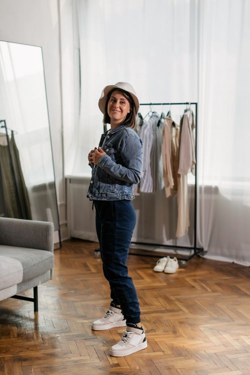 Free Young woman in a denim jacket and bucket hat posing stylishly indoors with clothing rack. Stock Photo