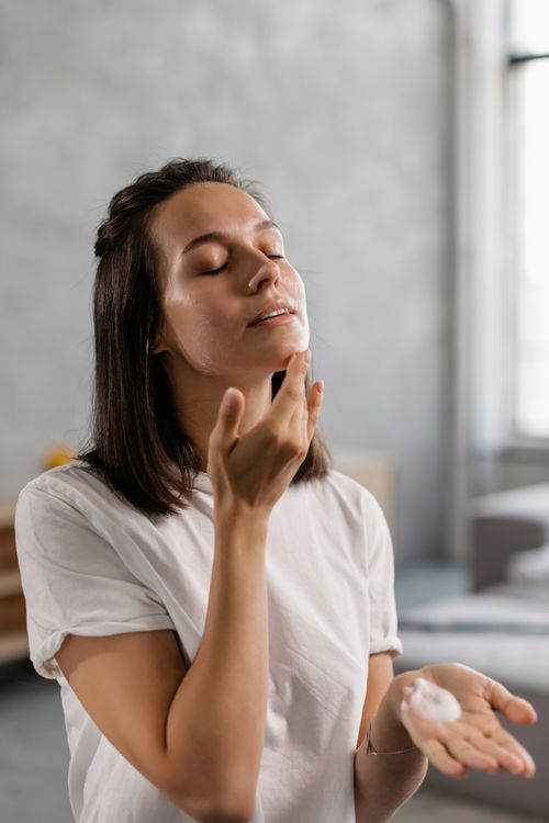 Free Young woman applying skincare cream to her face in a bright indoor setting. Stock Photo