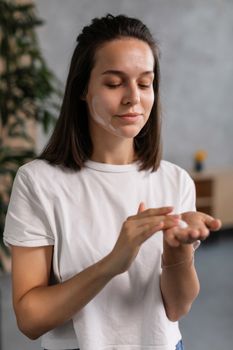 A young woman applies skincare cream to her hands indoors, promoting self-care.