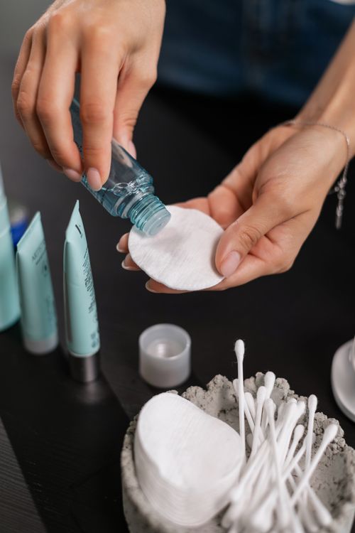 Close-up of hands applying toner to a cotton pad with skincare products in the background