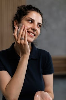 Woman smiling while applying facial cream indoors, exemplifying facial care routine.