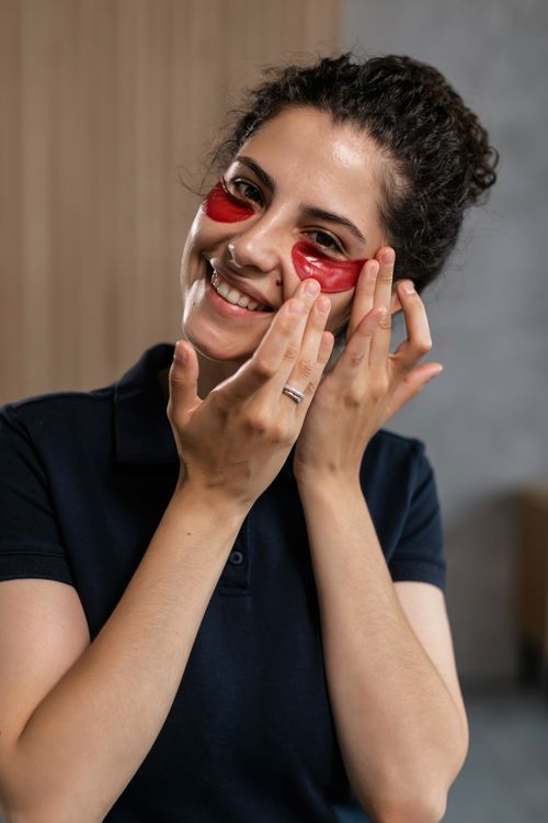 Free Happy woman applying red under-eye masks as part of her skincare routine indoors. Stock Photo