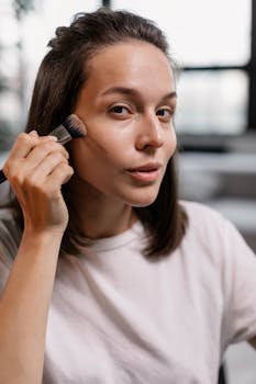 Young woman using a makeup brush on her face indoors, showcasing beauty routine.