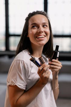 Happy woman indoors holding makeup brushes and products, showcasing beauty routine.