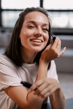 Smiling woman applying mascara indoors with natural lighting and a soft background.