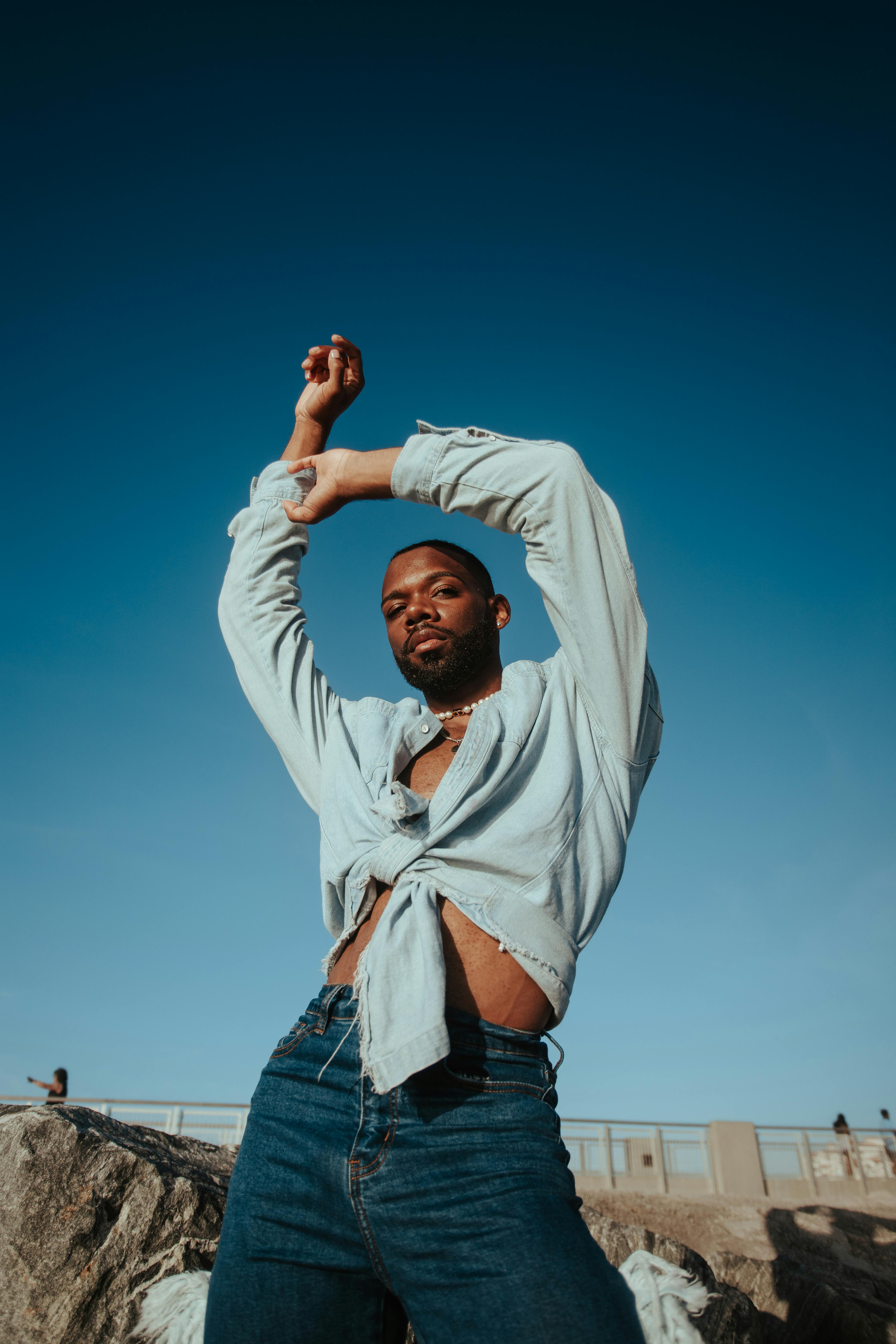 Fashion-forward pose of a young black man outdoors in a blue shirt and jeans under a clear summer sky.