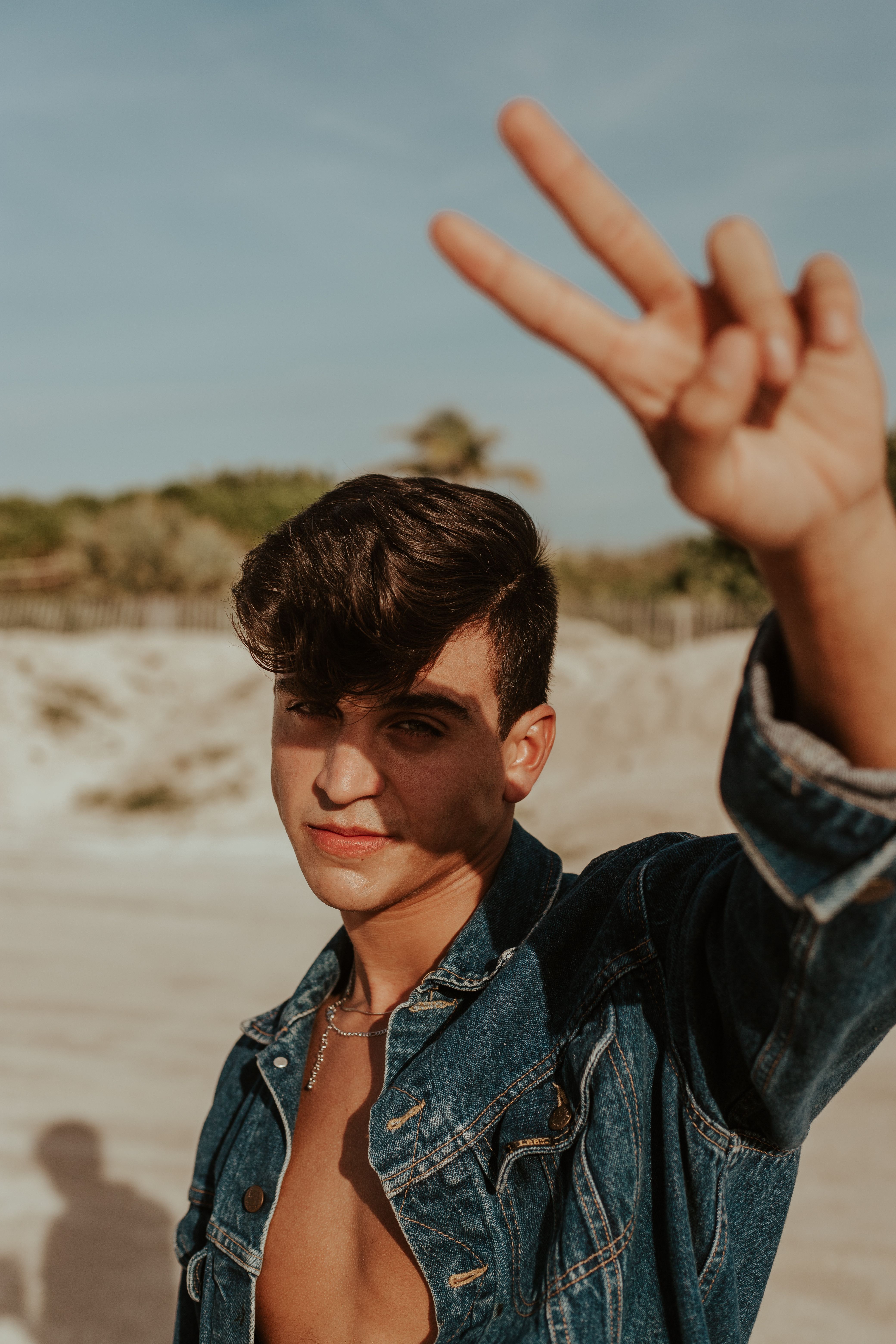 Stylish young man in a denim jacket flashing a peace sign at the beach on a sunny day.